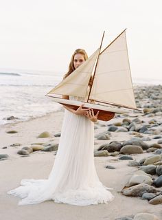 a woman holding a model sailboat on top of a sandy beach next to the ocean