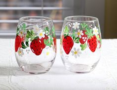 two wine glasses decorated with strawberries and daisies on a white tablecloth next to a window