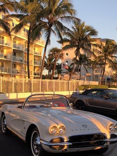 two classic cars parked next to each other in a parking lot with palm trees behind them