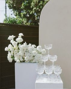 white flowers and wine goblets are arranged on a pedestal in front of a wall