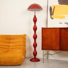 a red floor lamp sitting next to a yellow chair in a room with a painting on the wall