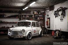 an old white mini car parked in a garage next to a shelf full of tools