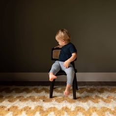 a little boy sitting on top of a black chair in front of a gray wall