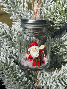 a christmas ornament in a mason jar hanging from a pine tree with snow on it