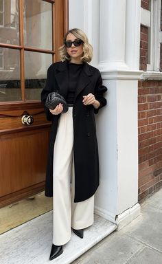 a woman in black coat and white pants leaning against a wall with her hand on her hip