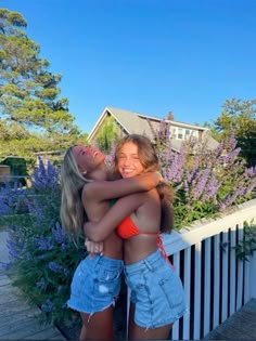 two young women hugging each other in front of purple flowers and a white picket fence