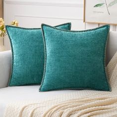 two teal pillows sitting on top of a white couch next to a vase with flowers