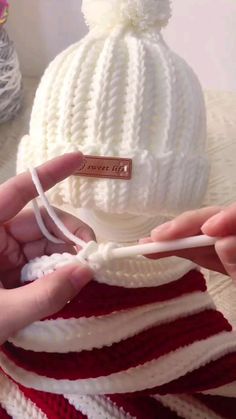 someone is knitting a white hat with red and white stripes on it, while another person holds the yarn in their hand