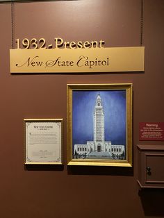 two framed pictures hang on the wall in front of a sign that reads, 1932 present new state capitol