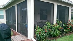 a screened in patio area with chairs and table next to plants on the side of the house