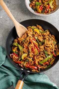 a skillet filled with noodles and vegetables on top of a blue towel next to a wooden spoon