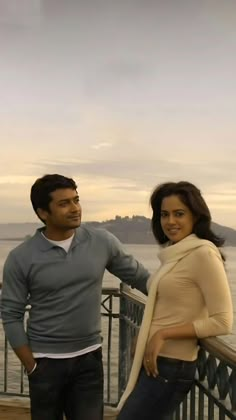 a man standing next to a woman on top of a bridge near the ocean with mountains in the background