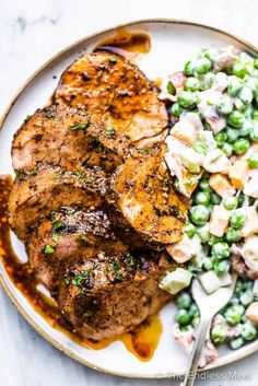 a white plate topped with meat, peas and salad next to a fork on a table