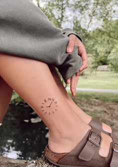 a woman's foot with a small compass tattoo on her left side leg, sitting next to a body of water