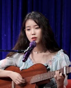 a woman holding a guitar and singing into a microphone while sitting in front of a blue curtain