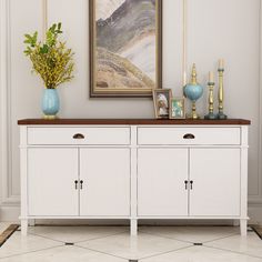 a white sideboard with three doors and two vases on the top, in front of a painting