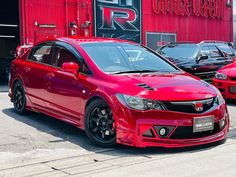 a red car is parked in front of a building with other cars and trucks behind it