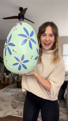 a woman is holding an easter egg in her hand and smiling at the camera while standing next to a ceiling fan