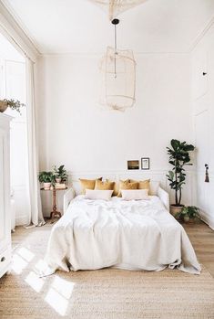 a white bed with yellow pillows in a bedroom next to a window and potted plants