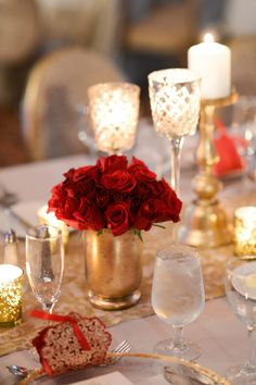 red roses in a gold vase on top of a table with candles and napkins