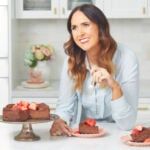 a woman sitting at a kitchen table with two cakes on the counter and another cake in front of her