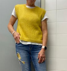 an older woman standing in front of a white wall wearing jeans and a yellow sweater