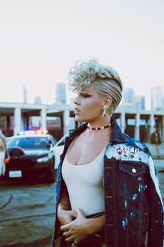 a woman with blonde hair wearing a jean jacket and necklace standing in front of a parking lot