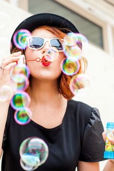 a woman blowing bubbles in the air while wearing sunglasses and a black hat with red lips