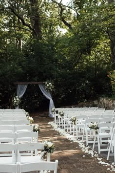 an outdoor wedding setup with white chairs and flowers on the aisle, surrounded by greenery