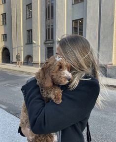 a woman holding a dog in her arms while walking down the street with buildings behind her