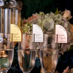 there are four wine glasses on the table with place cards in them and flowers behind them