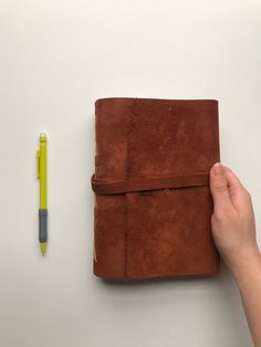 a hand holding a brown leather journal next to a yellow pen on a white surface