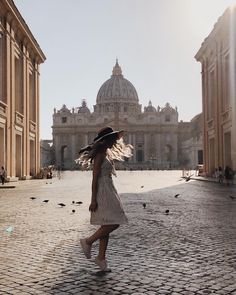 a woman in a dress and hat walking down a cobblestone street with pigeons on the ground