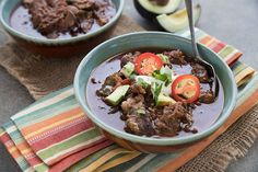 two bowls filled with beef and vegetables next to an avocado on a towel