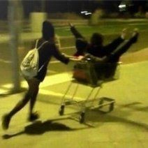 two people pushing a shopping cart down the street at night time with one person on it