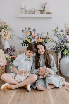 two young children are sitting on the floor with stuffed animals and flowers in the background