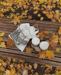 a newspaper and cups on a wooden bench in the fall leaves with someone's feet