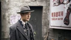 a man wearing a suit and hat standing in front of a brick wall with an advertisement on it