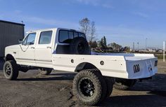 a white pickup truck parked in front of a black building with large tires on it's flatbed