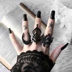 a woman's hand with black nail polish and rings on it, next to an open book