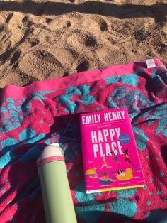 a pink book sitting on top of a beach towel next to a green cup and an umbrella