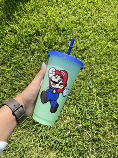 a hand holding a cup with an image of mario on it in front of some green grass