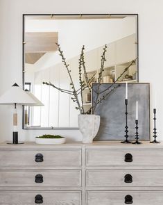 a white dresser topped with a vase filled with flowers next to a mirror and lamp