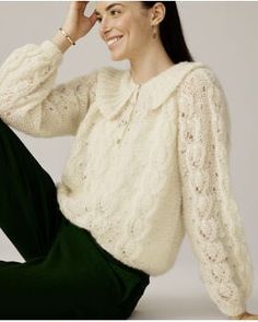 a woman sitting on the ground wearing green pants and a white sweater with ruffles