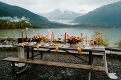 an outdoor table with candles and flowers on it near the water in front of mountains