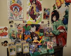 there are many anime pictures on the wall above the table and in front of them is an assortment of toys