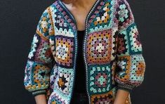 a woman standing in front of a black wall wearing a colorful crocheted jacket