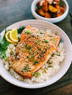 a white plate topped with salmon and rice next to a bowl of vegetables on a wooden table