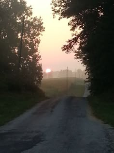 the sun is setting behind some trees on this dirt road with grass and bushes to the side