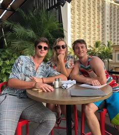 two men and a woman sitting at a table in front of a hotel with palm trees